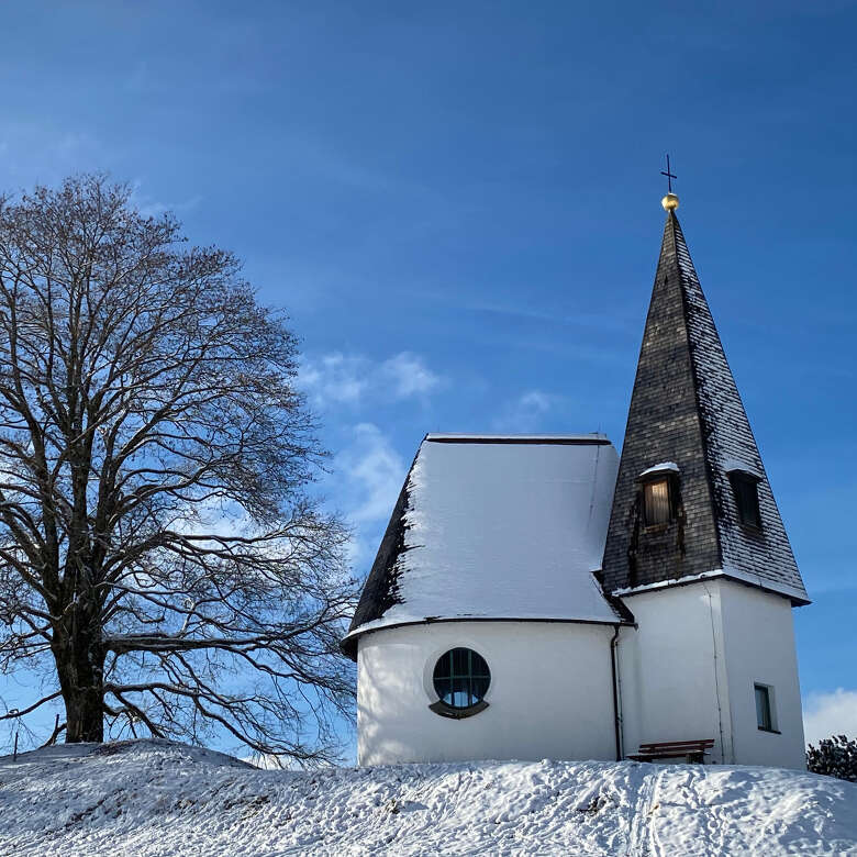 Alte Kapelle auf einer verschneiten Wiese, daneben ein kahler Baum.