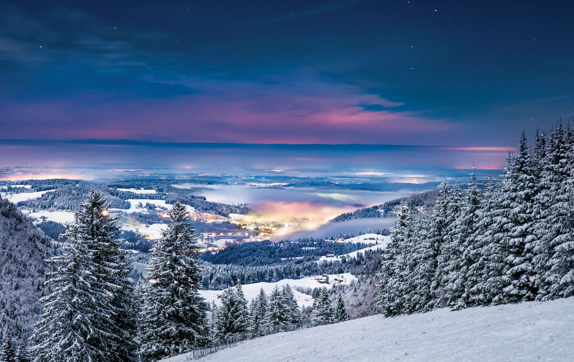 Der Nebel liegt im winterlichen Tal über Oberstaufen bei Nacht.