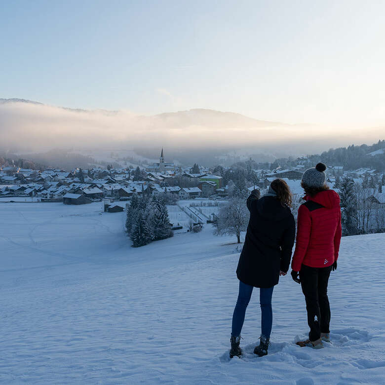 Winterwandern mit Blick auf das verschneite Oberstaufen mit Nebelstimmung im Sonnenuntergang.