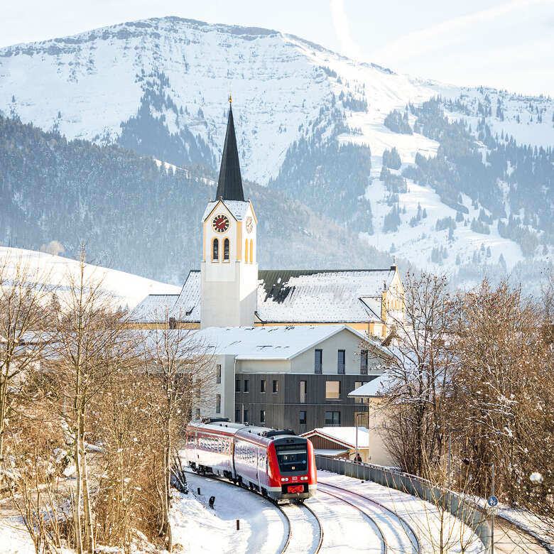 Zug fährt durch das Ortszentrum von Oberstaufen im Winter mit verschneiten Hochgratpanorama.