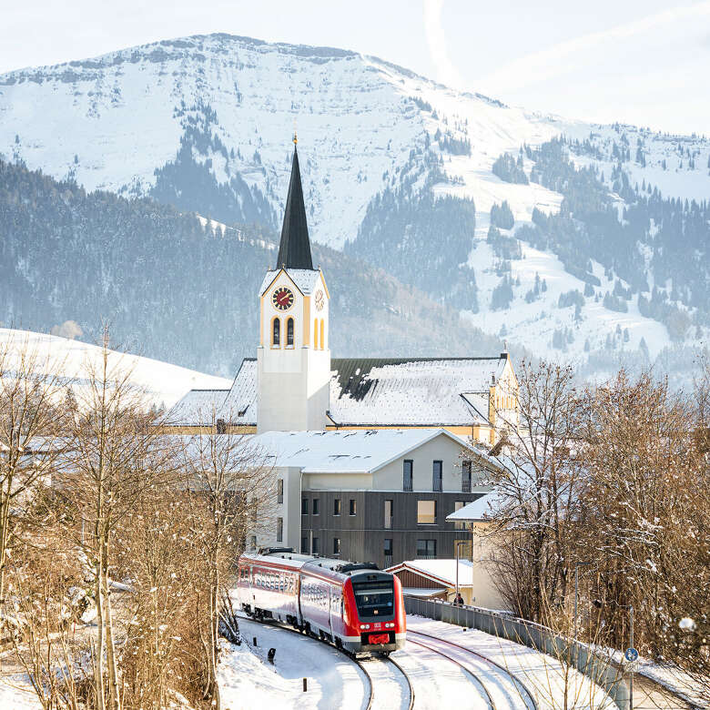 Zug fährt durch das Ortszentrum von Oberstaufen im Winter mit verschneiten Hochgratpanorama.