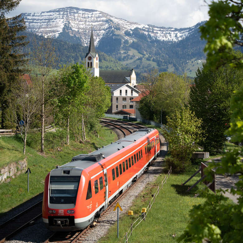 Zug auf der Bahnlinie durch Oberstaufen im Frühling mit dem verschneiten Hochgrat