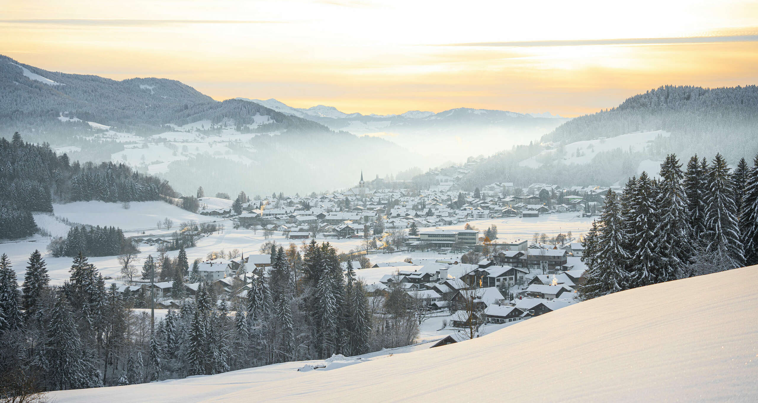 Ortsansicht von Oberstaufen in verschneiter Landschaft mit Bergpanorama im Sonnenuntergang.